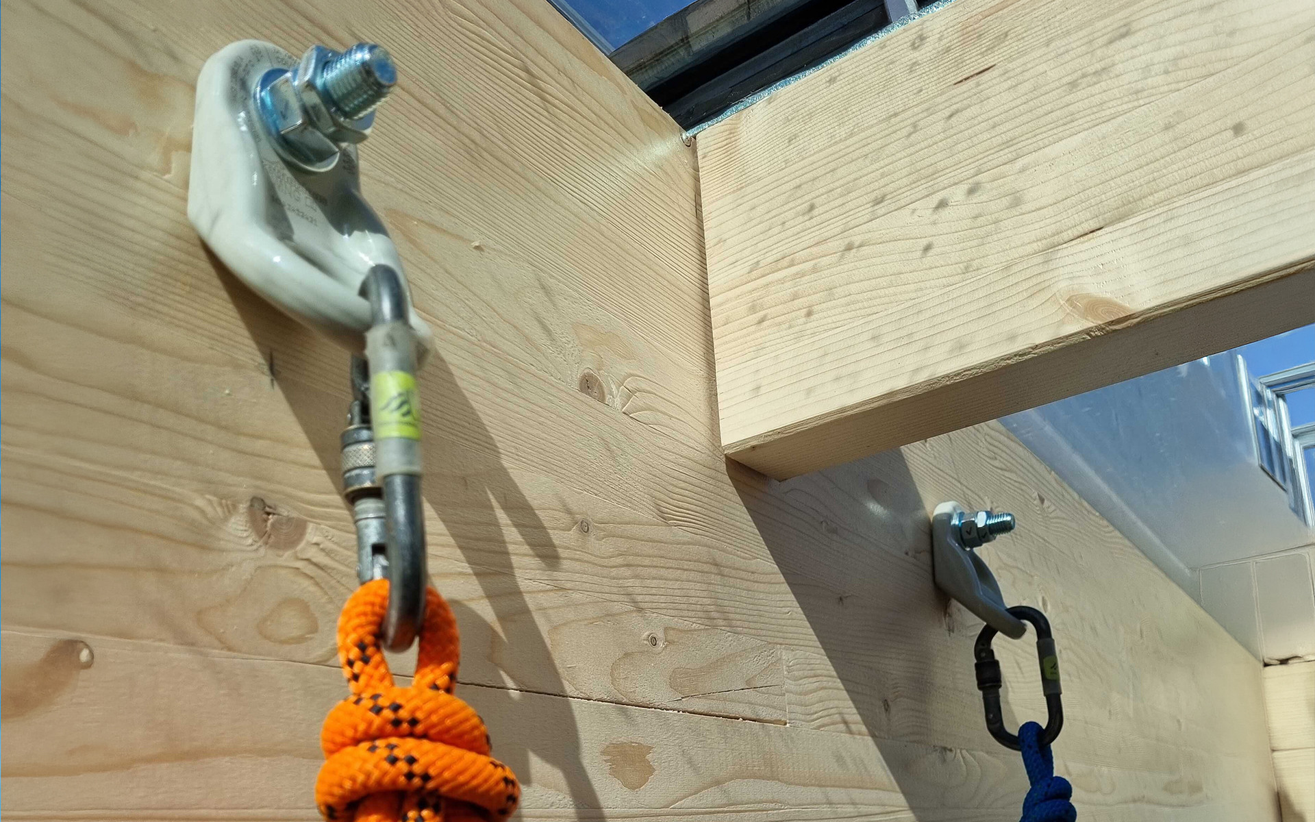 Rothoblaas WING single anchor points installed on a glulam timber beam junction — rope access carabiners and ropes connected for suspended work below a timber roof structure, Denmark.